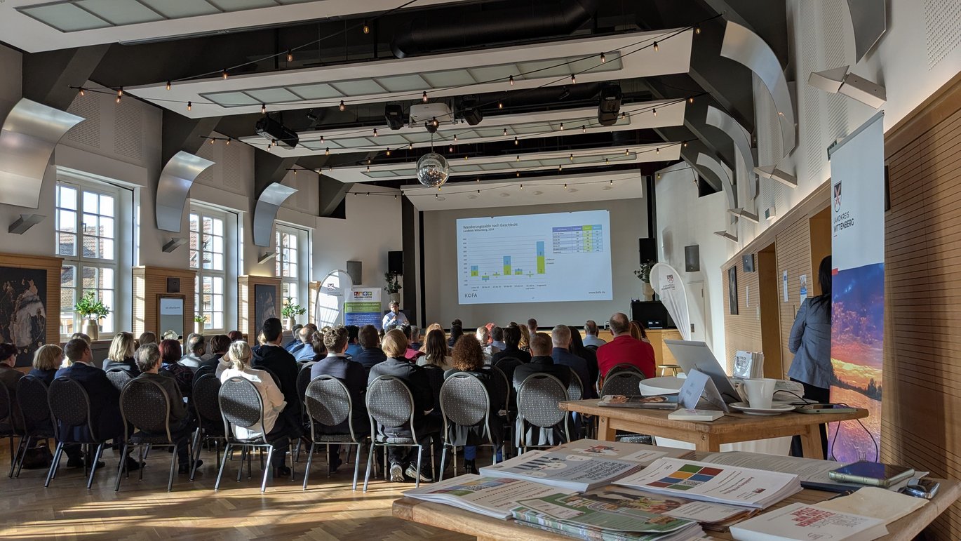 Publikum eines Vortrags von hinten, im Vordergrund ein Infostand mit Flyern und Broschüren