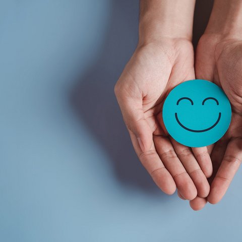 Ein lachender Smiley auf blauem Papier wird von zwei Händen präsentiert.