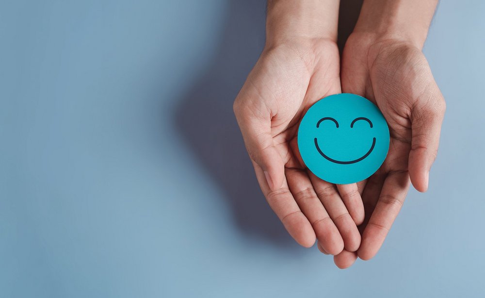 Ein lachender Smiley auf blauem Papier wird von zwei Händen präsentiert.