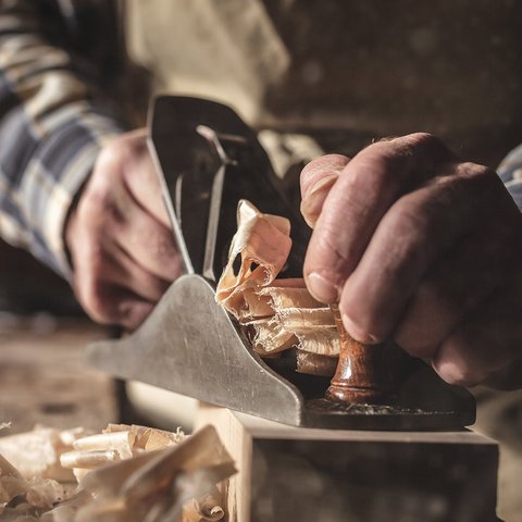 Zu sehen sind zwei Männerhände die mit einem alten Hobel ein großes Stück Holz bearbeiten.